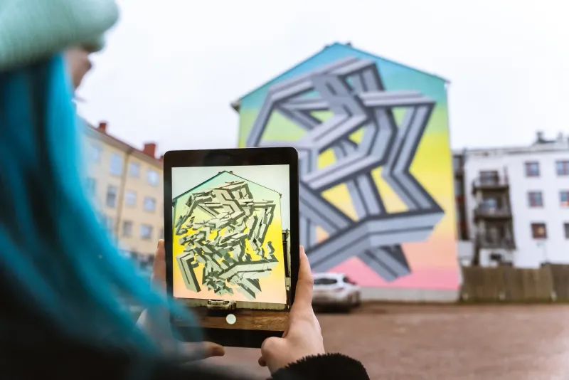 A tablet held up to a geometric street mural, showing the artwork lifting off the wall in 3D